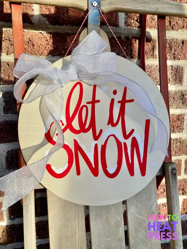 round wooden sign with red and white letters saying 'let it snow' with white and silver bow